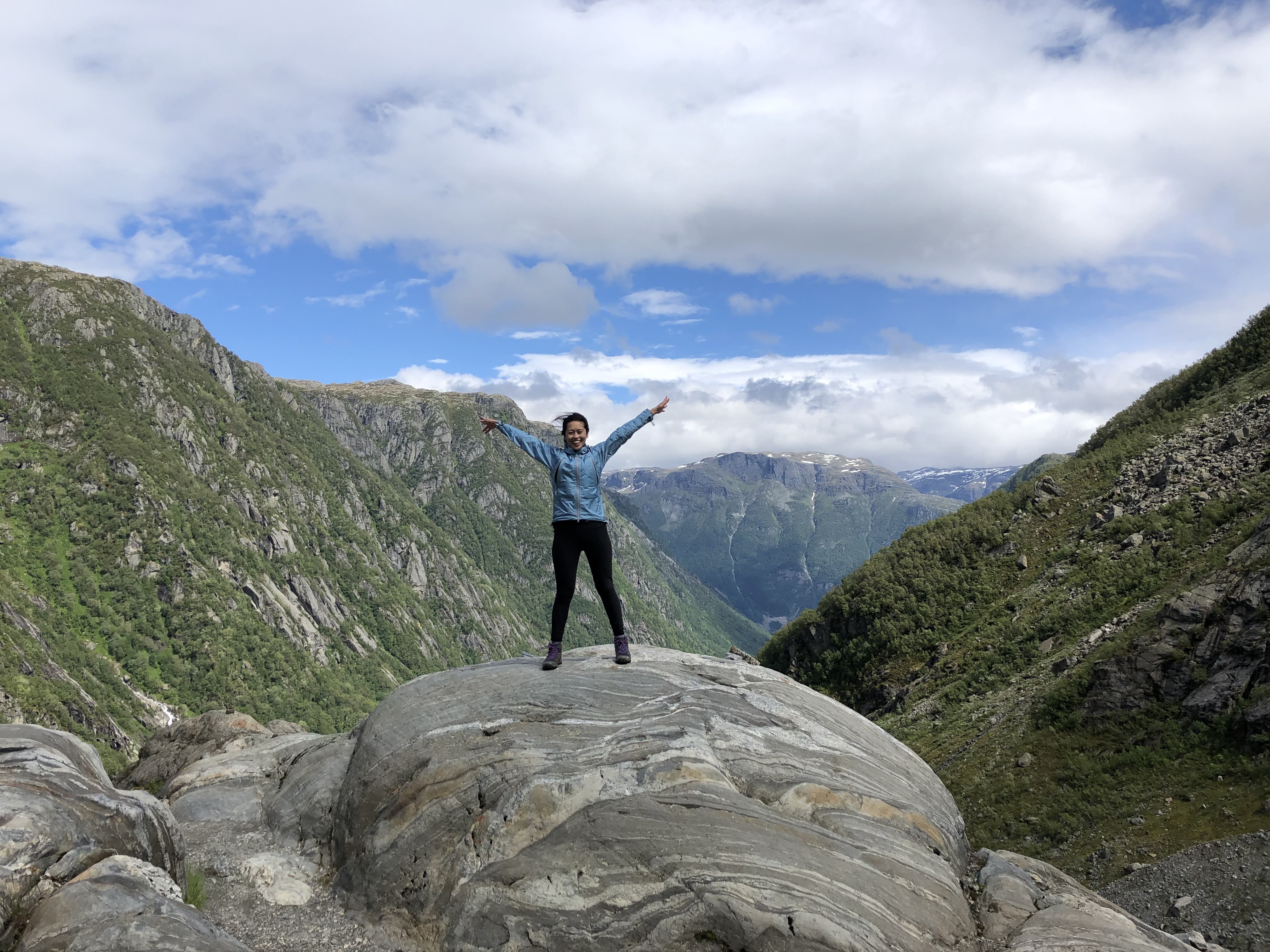Cindy stands in a star position atop a huge rock in the middle of the mountains, smiling tains