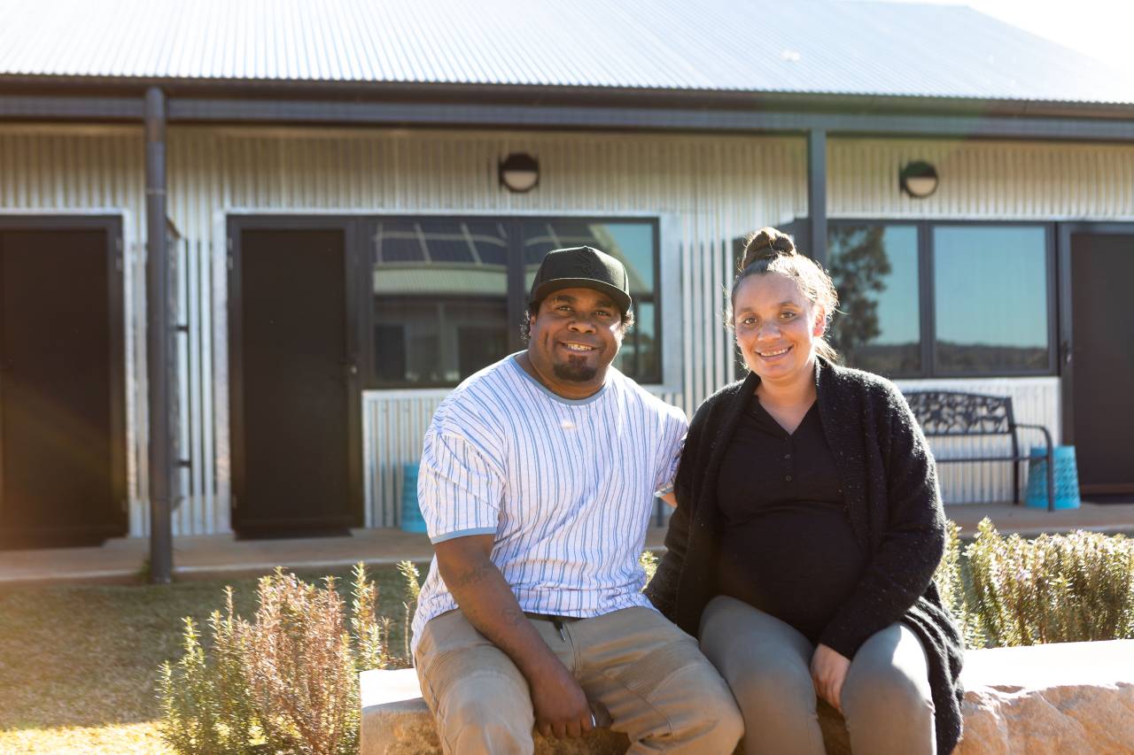 A man wearing a cap and a woman wearing a cardigan sitting on a ledge, smiling