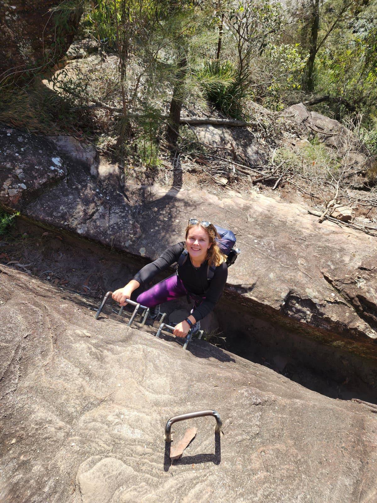 A smiling woman with blonde hair climbing up a rock face carrying a backpack