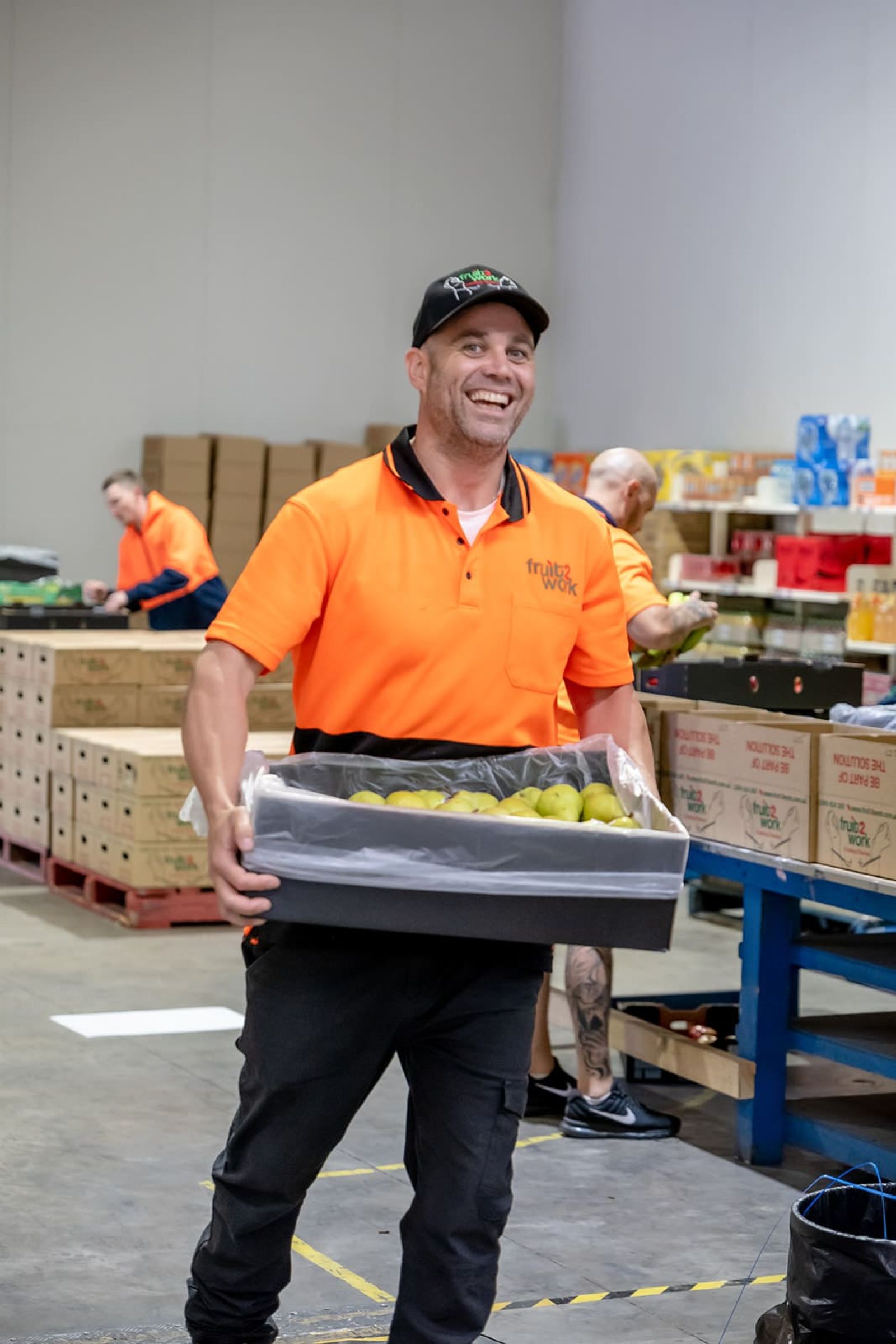 A man in an orange shirt and black hat carries a box of fruit