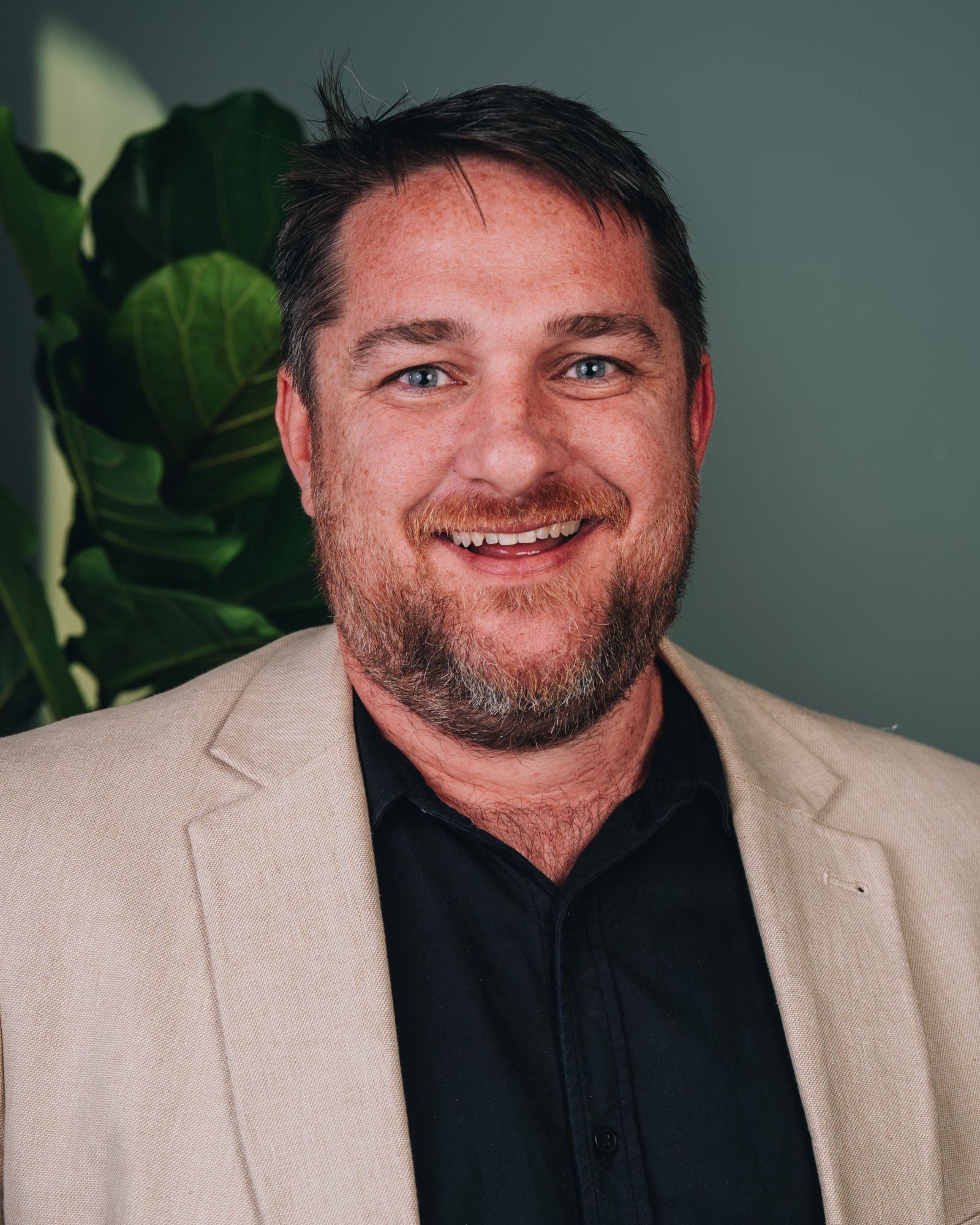 Joel wears a beige blazer on top of a black shirt with a big smile