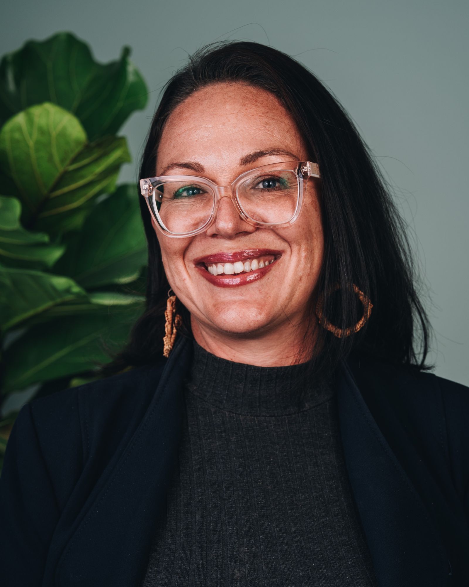 Chelsea is smiling, wearing glasses and large circular earrings and a suit jacket